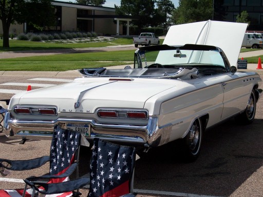 White 1960s Buick convertible with top down and hood up at a car show, rear three-quarter view