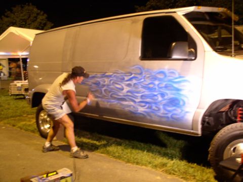 1978 Ford Mustang II - Artist airbrushes blue flames onto the side of a van during an outdoor event.