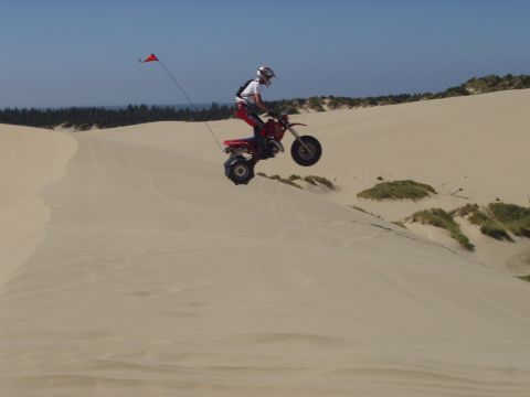 1991 Ford Bronco - A rider on a red three-wheeled ATV soars through the air while jumping a sand dune.