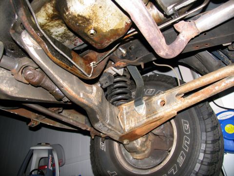 1984 Ford Bronco - Underbody shot from beneath a lifted truck showing the rusty Twin Traction Beam front suspension: axle beam, radius arm, coil spring with a…