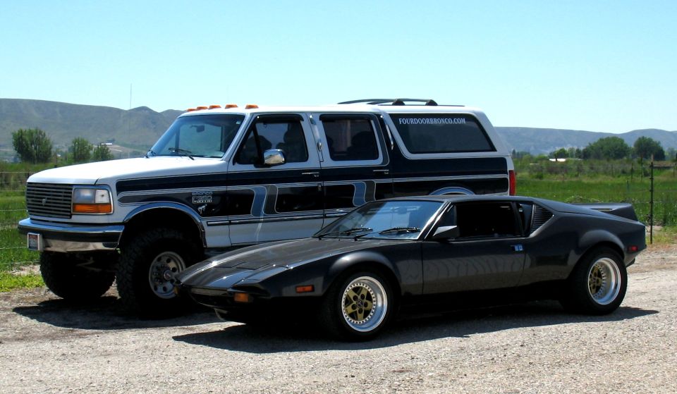 Charcoal DeTomaso Pantera coupe parked beside a black-and-white four-door Bronco on gravel.