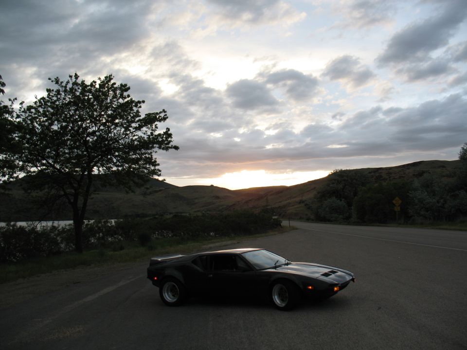 Black DeTomaso Pantera coupe parked on roadside at sunset near hills and lake