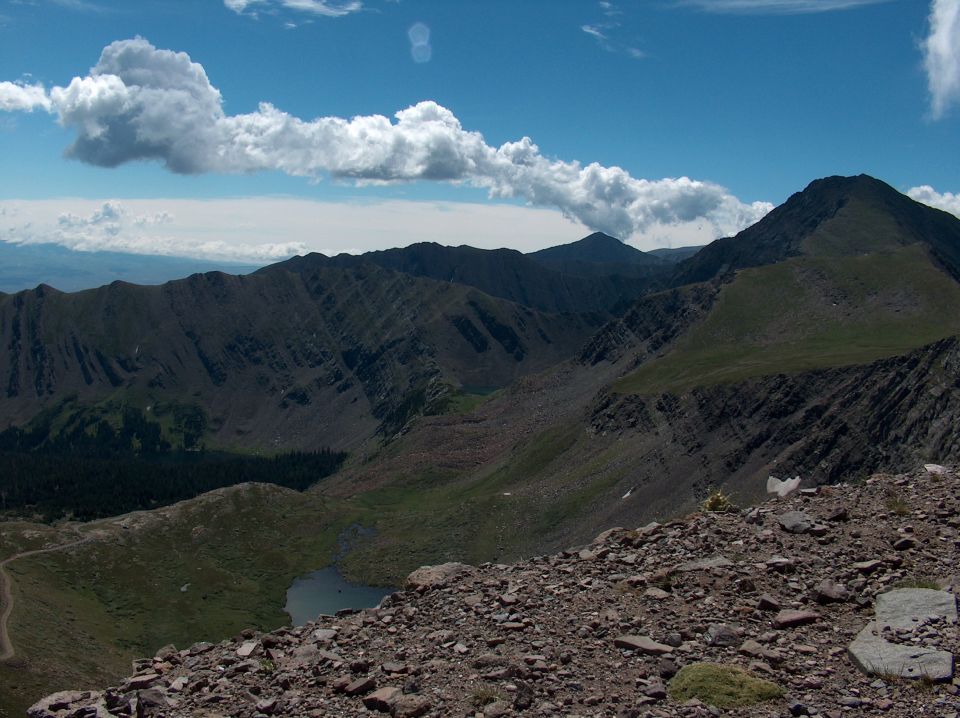 Mountain range under a blue sky with white clouds, rocky foreground, small lake in valley