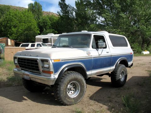 1979 Ford Bronco - A two-tone white and blue Ford Bronco is parked outdoors with other vehicles and structures in the background.