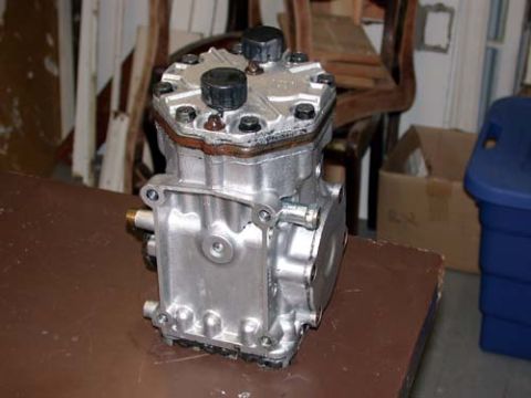 1996 Ford Bronco - A cast-aluminum York-style piston compressor sitting on a brown workbench in a cluttered garage, with two black service port caps and hex h…