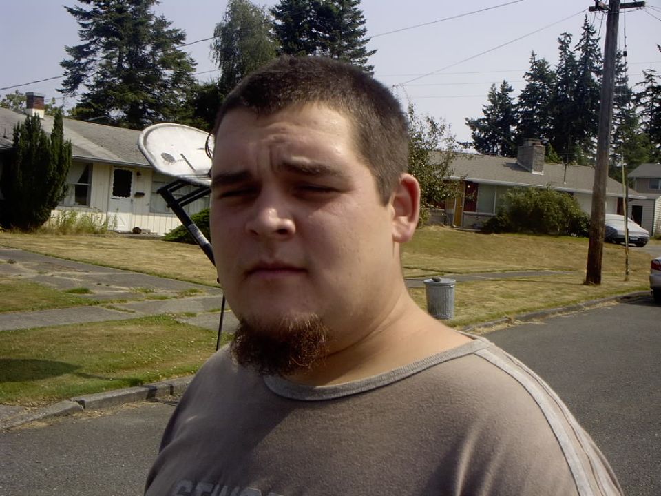 Young man with goatee in gray shirt standing on a suburban street; no vehicle in focus