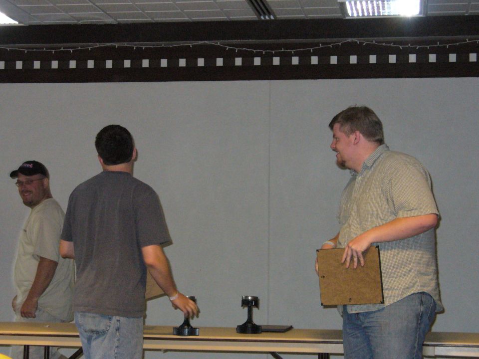 Three men stand behind a table with trophies; one man on the left wears a baseball cap