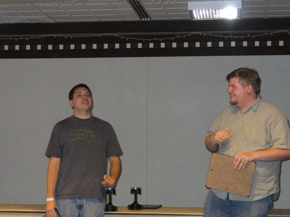 Two men standing indoors, one wearing a grey t-shirt with text, the other holding a clipboard