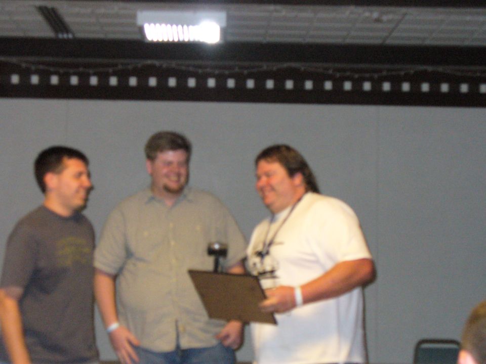 Three men stand in front of a gray wall, one holding a plaque