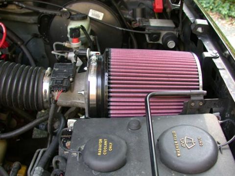 1996 Ford Bronco - Engine-bay close-up of a red K&N-style conical cone air filter clamped to a chrome intake tube, replacing the factory airbox, with the mass…