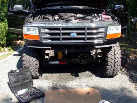 1996 Ford Bronco - Head-on view of a black 1990s Ford Bronco in a driveway with the hood up and the front bumper removed, exposing the bumper mounting bracket…