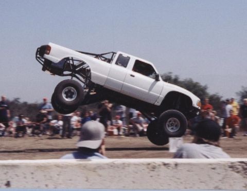 1984 Ford Bronco - A white pickup truck performs a jump, its front wheels lifted high off the ground.