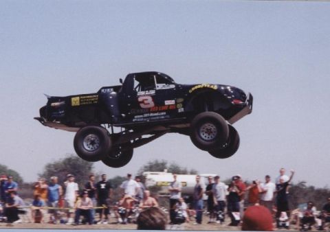 1984 Ford Bronco - A custom off-road truck takes flight during a race event, with spectators watching from the sidelines.