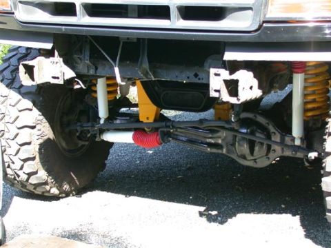 1996 Ford Bronco - Underside view of a lifted Ford truck front suspension shot from ground level, showing a solid front axle with orange coil springs, a red-b…