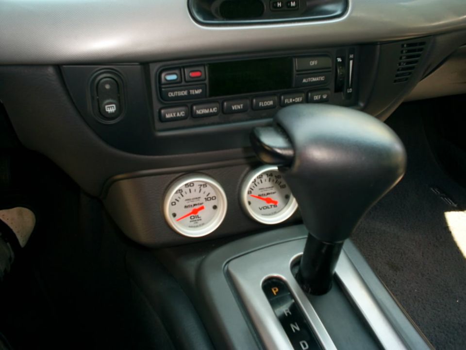 Car interior showing automatic shifter, climate controls, and two aftermarket gauges for oil and voltage