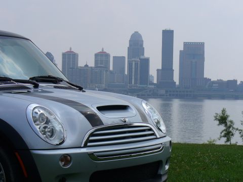 1991 Ford Probe - A silver Mini Cooper is parked on a grassy hill overlooking a river and a city skyline.