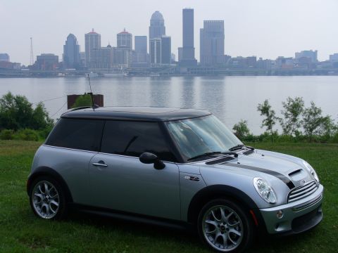 1991 Ford Probe - A silver Mini Cooper is parked on a grassy hill overlooking a river and a city skyline.