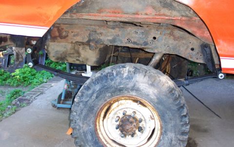 1978 Ford Bronco - Close-up view of the rear wheel and suspension system of an orange vehicle, highlighting its modified leaf springs and rusty undercarriage.