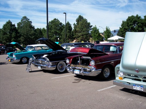 Several classic cars parked in a lot with hoods up. One is teal, one is black, one is maroon, and a light blue truck is in the foreground