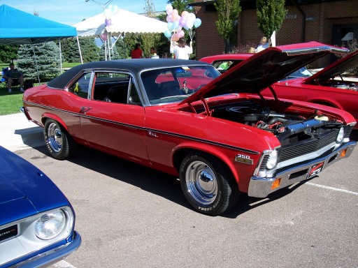 Red coupe with black vinyl top, hood open showing engine. Another red car behind it. Blue car in foreground