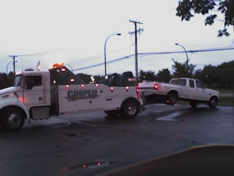 1992 Ford Bronco - A white pickup truck is being towed by a tow truck on a wet road under an overcast sky.