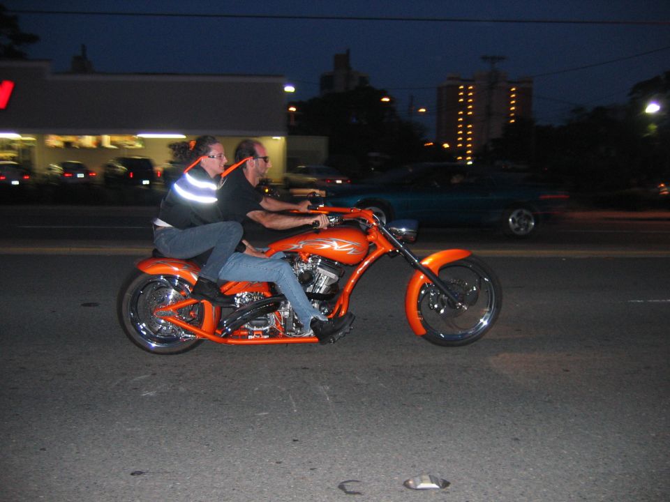 Two people ride a bright orange custom motorcycle down a street at dusk