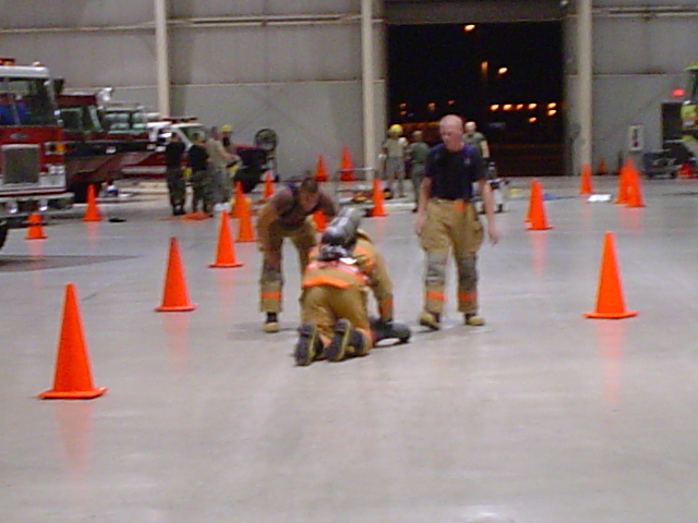 Firefighters in gear practice around orange cones inside a large building, with fire trucks visible in the background