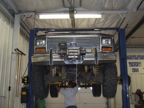 1986 Ford Bronco - A black Ford Bronco is raised on a hydraulic lift in a garage, with a mechanic working on its undercarriage.