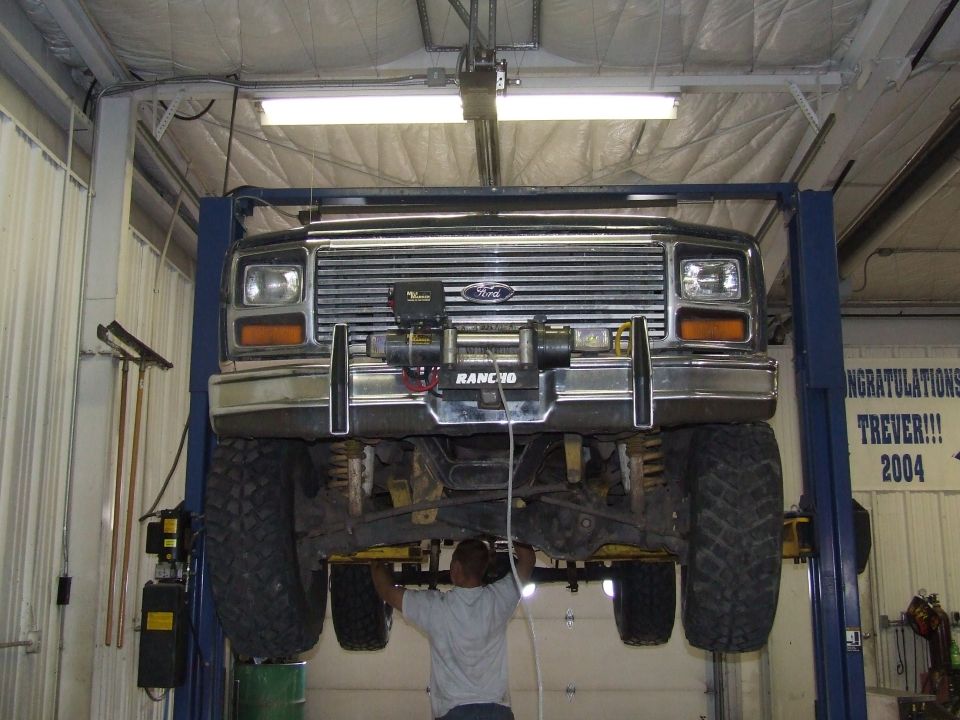 Black Ford Bronco on a lift, front view. Chrome grille, winch, and bumper. Large tires. Mechanic underneath