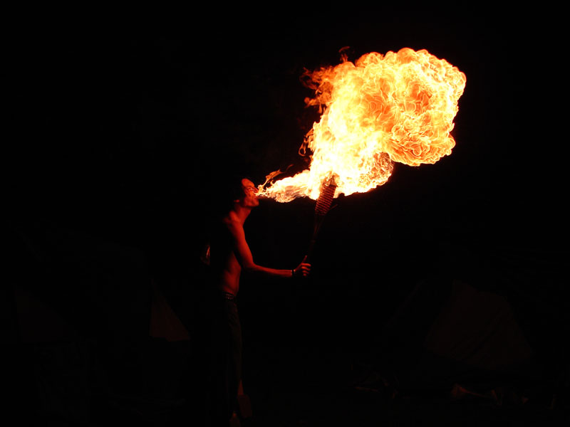 Person breathing a large plume of fire in the dark, holding a torch