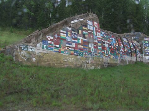 1990 Ford Bronco - A large concrete structure is decorated with numerous painted flags and graffiti, set against a backdrop of trees.