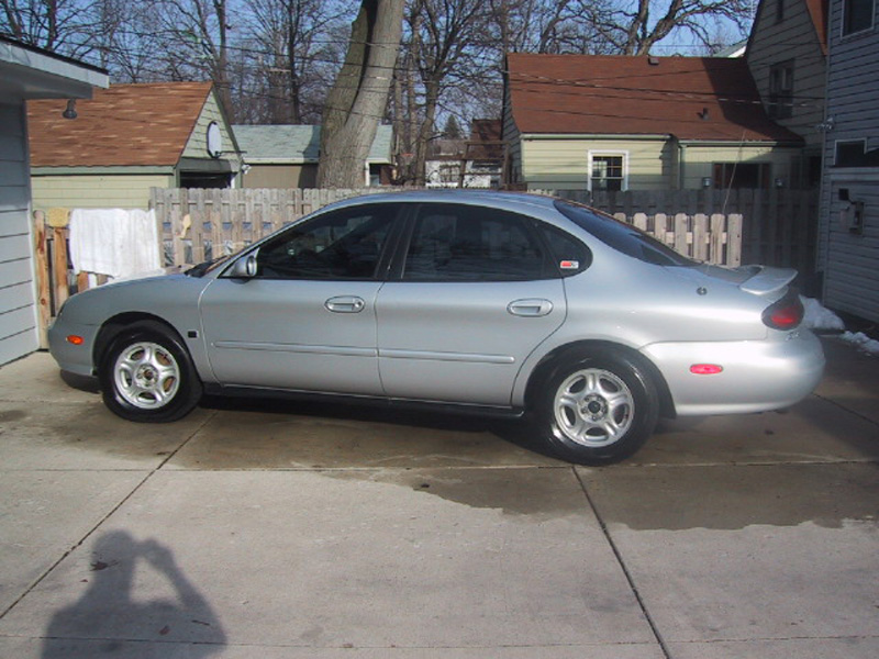 Silver Ford Taurus sedan, right side view, parked on a driveway with a wooden fence behind it