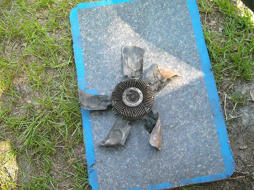 Damaged engine cooling fan with bent blades, resting on a blue-edged mat outdoors