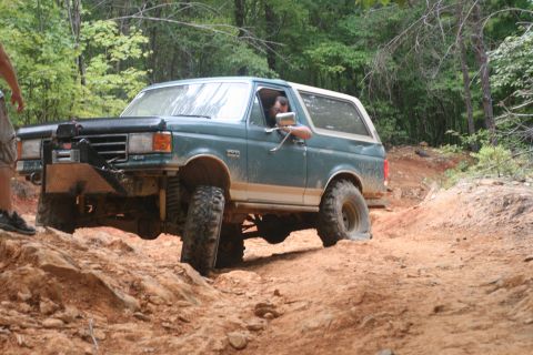 1990 Ford Bronco - A green Ford Bronco is shown off-roading on a dirt and rock path with trees in the background.