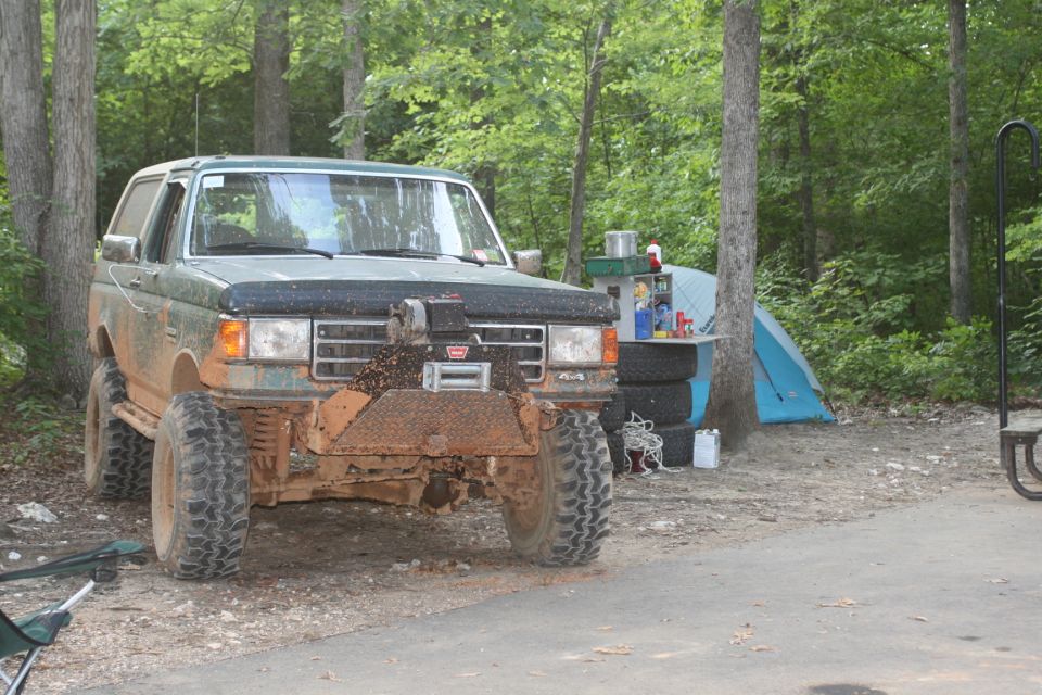 Mud-covered green Ford SUV with winch bumper parked at a wooded campsite with tent