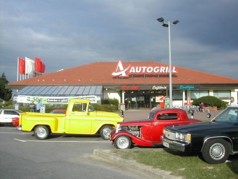 1991 Ford Crown Victoria - A trio of classic vehicles are parked outside an Autogrill restaurant under a cloudy sky.