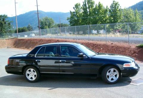 2002 Ford Crown Victoria - A black 2002 Ford Crown Victoria is parked on a paved surface with a chain-link fence and trees behind it.