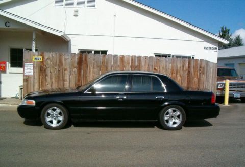 2002 Ford Crown Victoria - A black 2002 Ford Crown Victoria is parked next to a wooden fence, with a brown pickup truck visible in the background.