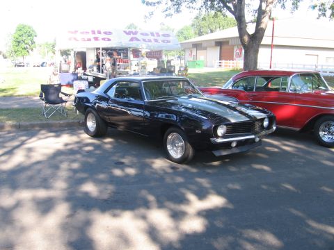 1989 Ford Mustang - A dark blue 1969 Chevrolet Camaro is parked next to a red classic car, with a vendor tent in the background.