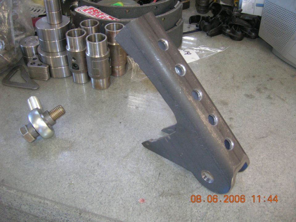 Bare steel multi-hole suspension link bracket on shop floor beside machined bungs and a rod end