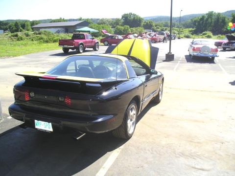 1984 Pontiac Fiero - Black 1990s Pontiac Firebird Trans Am with yellow hood stripes and a custom plate is parked at an outdoor car show.
