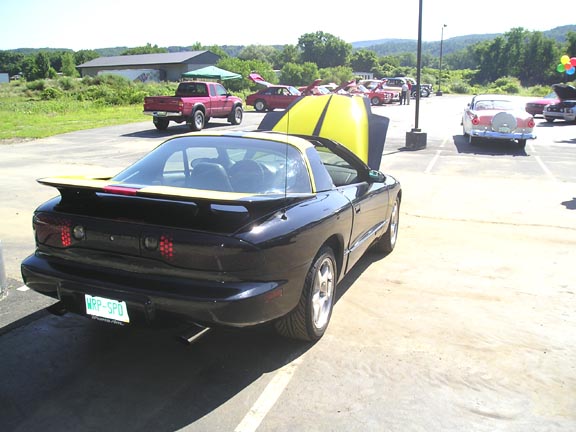 Black 1990s Pontiac Firebird Trans Am with yellow hood stripes, rear spoiler, and custom plate WRR-SPD. Other cars and a red truck are parked nearby