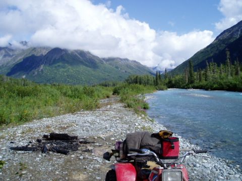 2000 Ford E350 - An ATV is parked on a rocky shore next to a river, with mountains and clouds overhead.