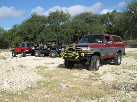 1995 Ford Bronco - A group of off-road vehicles, including a Ford Bronco and Jeeps, are parked in a dry, grassy area under a blue sky.