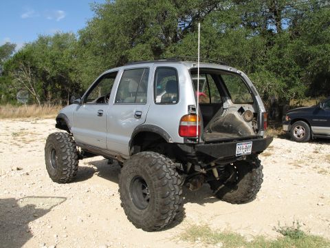 1995 Ford Bronco - Modified SUV with massive tires and lifted suspension sits in a dirt lot with trees in the background.