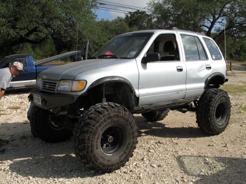1995 Ford Bronco - A heavily modified silver SUV with large off-road tires is parked on a gravel lot next to a blue pickup truck.