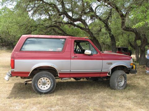 1995 Ford Bronco - A red and silver 1995 Ford Bronco is parked outdoors with another vehicle visible in the background.