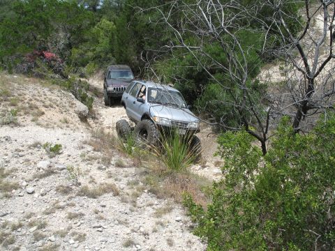 1995 Ford Bronco - Two SUVs traverse a challenging off-road path through a wooded, rocky landscape.