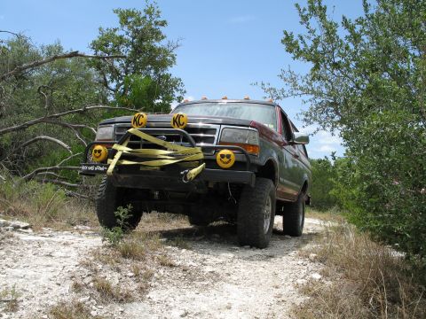 1995 Ford Bronco - This modified Ford Bronco is driving on a dirt road surrounded by trees and brush under a clear blue sky.