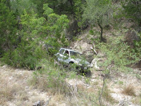 1995 Ford Bronco - A silver 1995 Ford Bronco is seen off-road, partially hidden by dense foliage on a rocky slope.